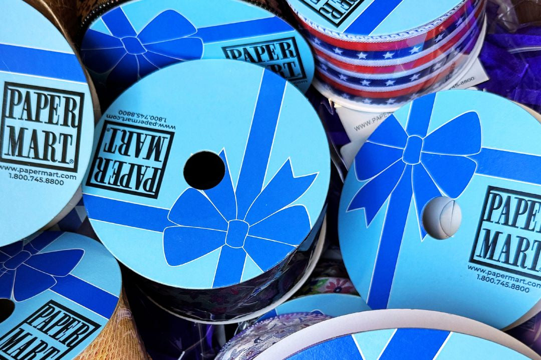 A close-up of multiple spools of ribbon with blue and white gift box-ribbon style labels reading PAPER MART, alongside stacks of red, white, and blue patterned ribbons.